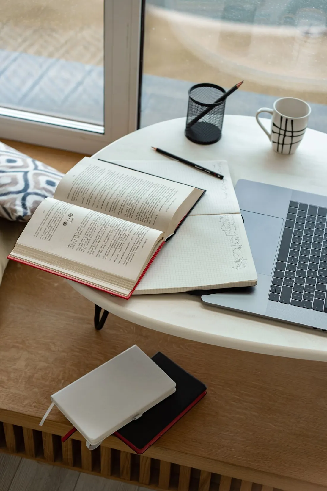 A notebook, a pencil and a mug beside a laptop on a small round table by a window