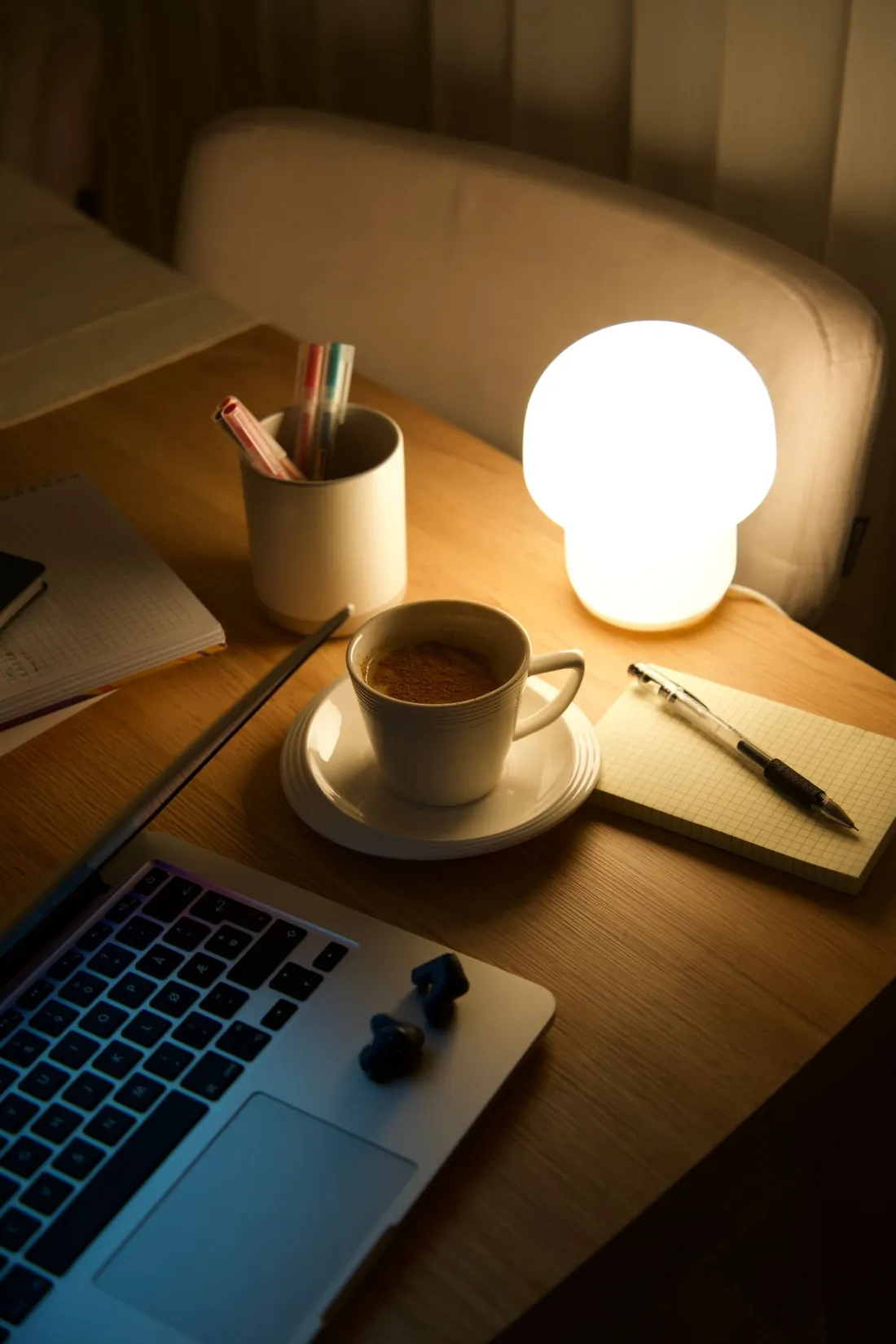 A laptop, a cup and a notepad under a small lamp on a desk at night
